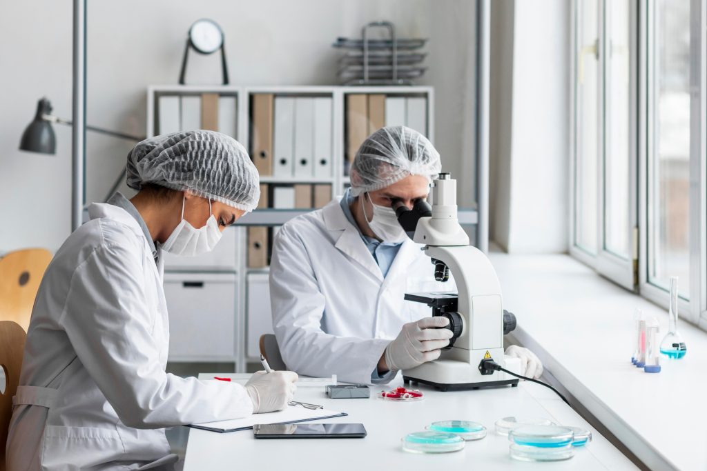 Dos investigadoras con mascarilla y gorro trabajando con un microscopio en un laboratorio